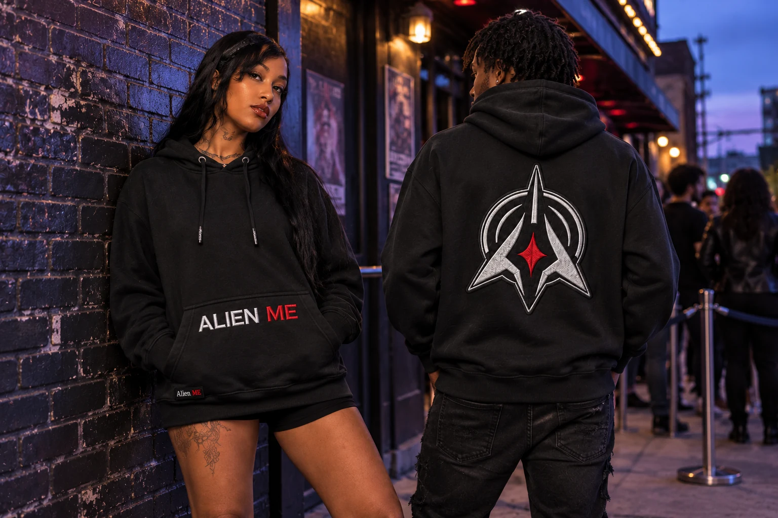 Two adult models wearing the black AM Emblem Hoodie outside a music venue at dusk, showing its pocket embroidery and back patch