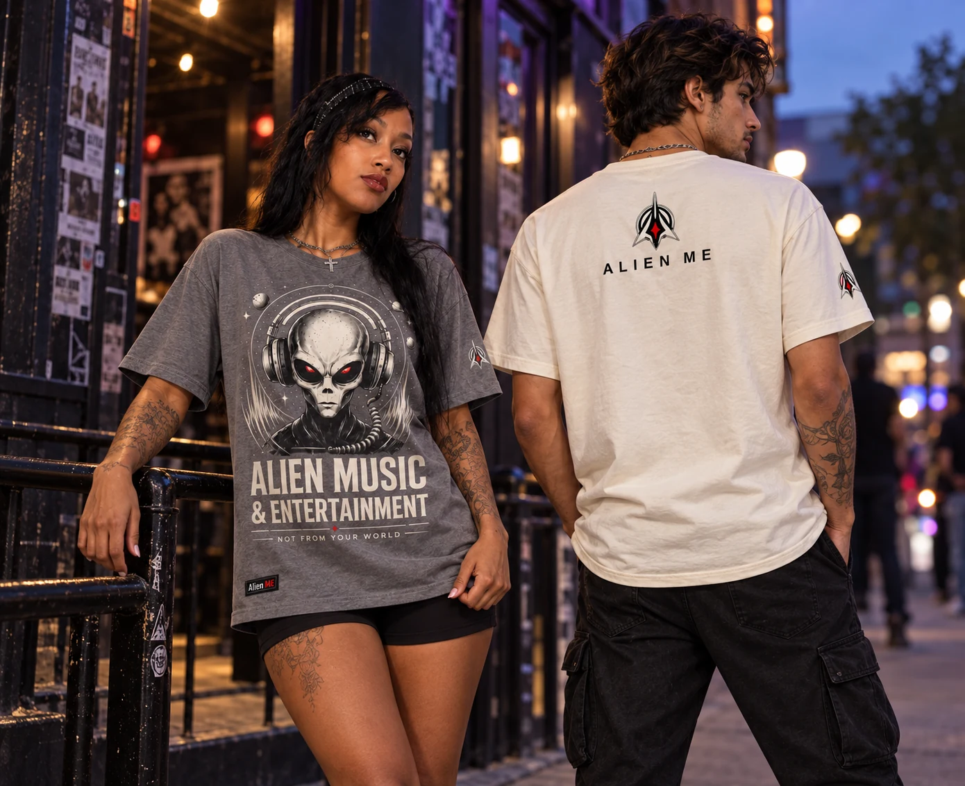 Two adult models wearing the Signature Alien Tee in gray and cream outside a music venue at dusk, showing front and back
